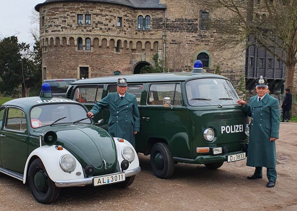 - Die Polizeikomparsen Eberhard Dersch (rechts) und Klaus Krawatzki mit dem VW Käfer bzw. VW T2 aus dem Marburger Museum beim Filmdreh zur ARD-Serie „Unsere wunderbaren Jahre“ vor dem Schloss Hülchrath